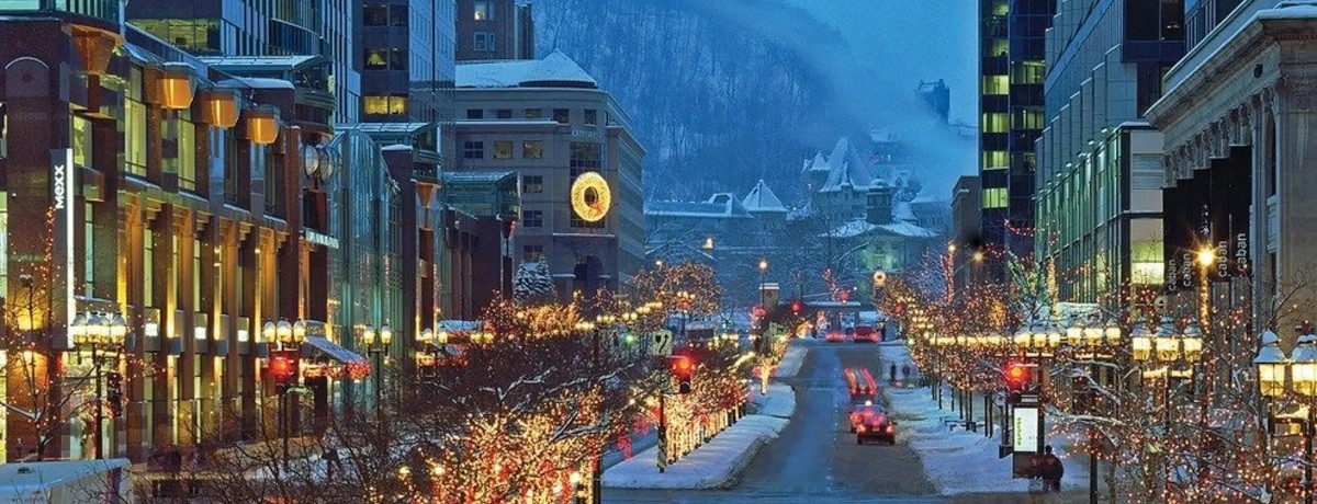Winter view of McGill College Avenue in Montreal with festive Christmas lights lining the street, cars and pedestrians visible, and Mount Royal in the background. Vue hivernale de l’avenue McGill College à Montréal avec des lumières de Noël décoratives le long de la rue, des voitures et des passants visibles, et le mont Royal en arrière-plan.