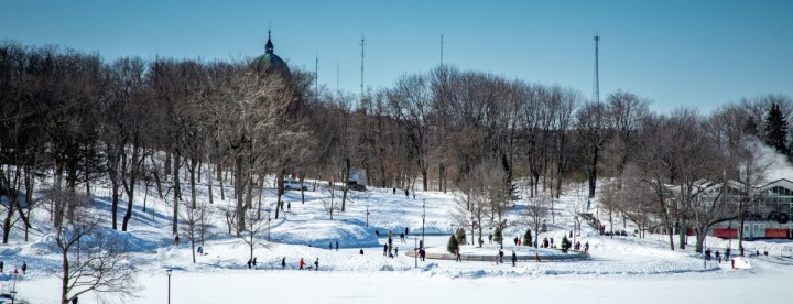 People skating outdoors on Beaver Lake rink in Montreal with snow-covered trees and Mount Royal in the background – Des personnes patinent sur la patinoire extérieure du Lac aux Castors à Montréal avec des arbres enneigés et le Mont Royal en arrière-plan.
