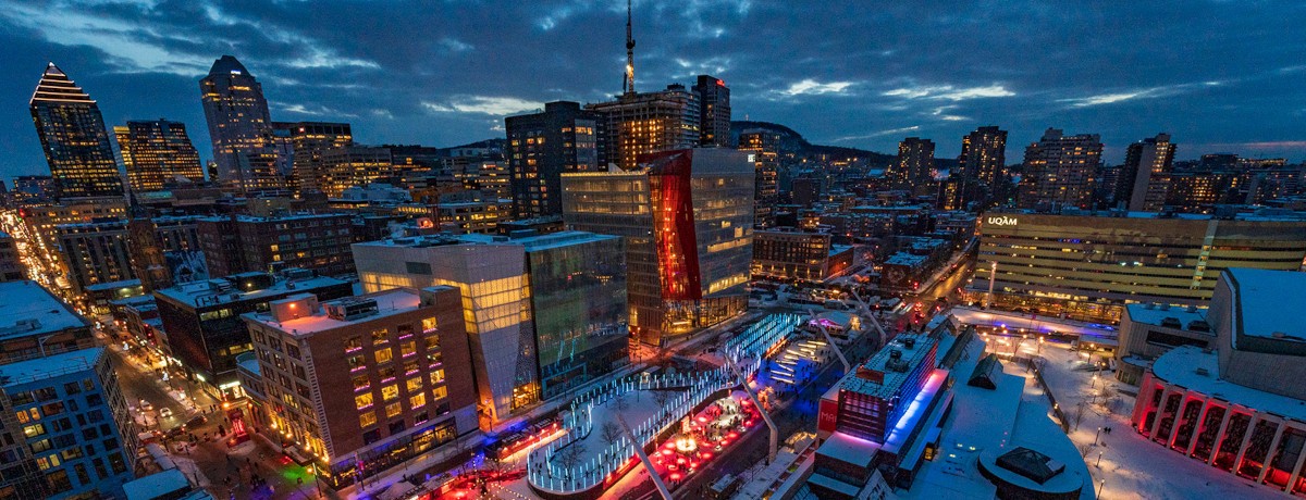 Aerial view of a vibrant city, Montreal, at night, showcasing brightly lit buildings and streets with colorful lights. Snow covers the ground, conveying a festive and lively winter atmosphere. | Vue aérienne nocturne de Montréal, ville animée où se dévoilent des bâtiments et des rues illuminés de mille feux. La neige recouvre le sol, créant une ambiance hivernale festive et chaleureuse.