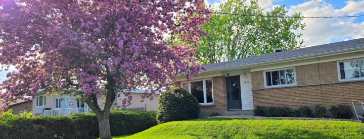 A charming brick house with a lush green lawn and a blooming pink tree on a sunny day. The sky is partly cloudy, creating a serene, inviting atmosphere. | Une charmante maison en briques, entourée d'une pelouse verdoyante et d'un arbre rose en fleurs, par une journée ensoleillée. Le ciel partiellement nuageux crée une atmosphère sereine et accueillante.