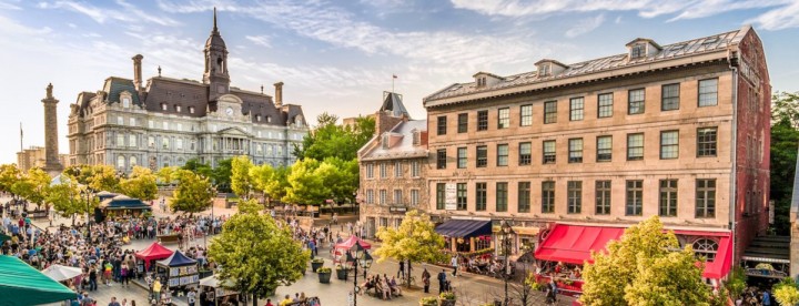 A lively street market scene with vibrant tents and many people milling around. Historic buildings under a blue sky create a bustling and cheerful atmosphere. | Une scène de marché de rue animée, avec ses tentes colorées et une foule dense qui s'y presse. Des bâtiments historiques sous un ciel bleu créent une atmosphère joyeuse et animée.